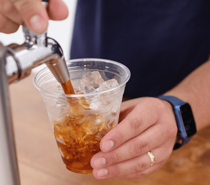 Person pouring a cold brew drink into a clear plastic cup with ice cubes on a wooden surface.