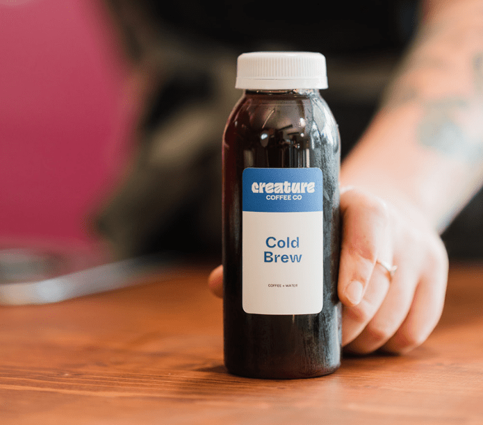 Bottle of creature Coffee Co. Cold Brew held by a person on a wooden surface.