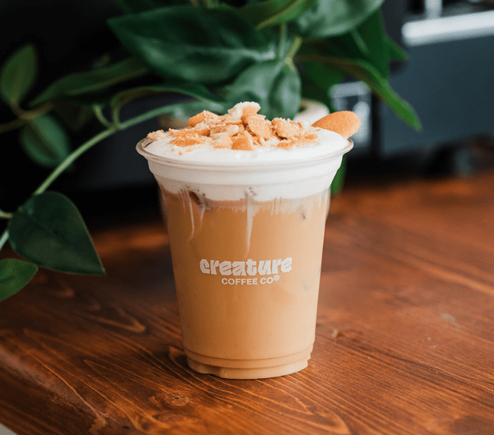 Coffee drink with whipped cream and cookies in a clear cup on a wooden surface, featuring 'creature coffee co' branding.