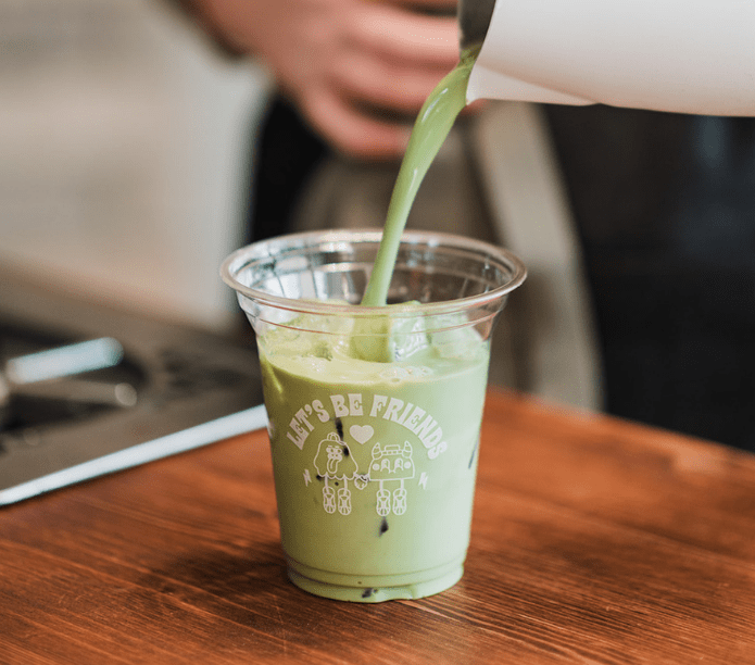 Matcha Latte being poured into a clear plastic cup with text on a wooden surface.