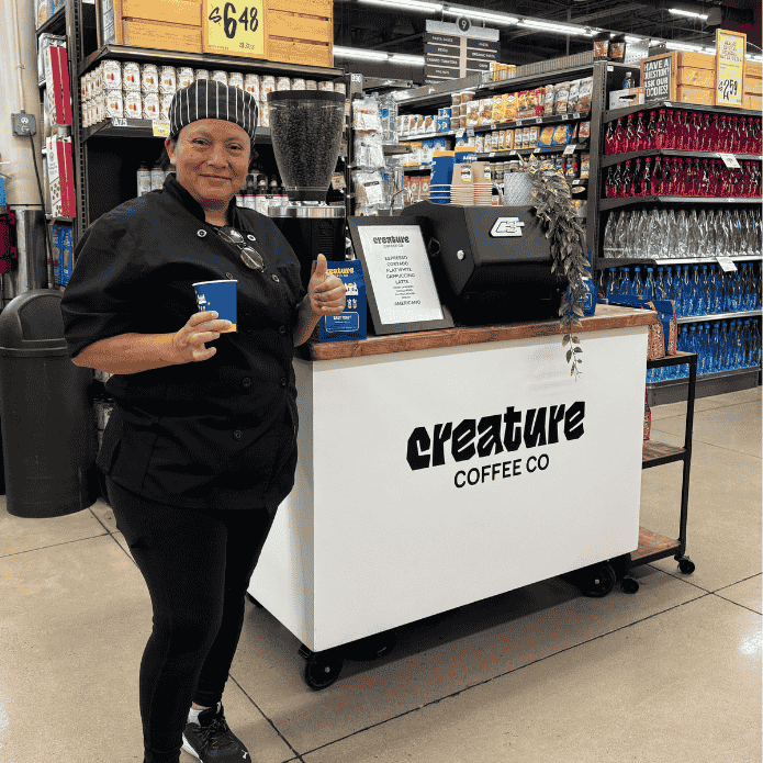Person standing next to a 'Creature Coffee Co.' coffee station in a store.