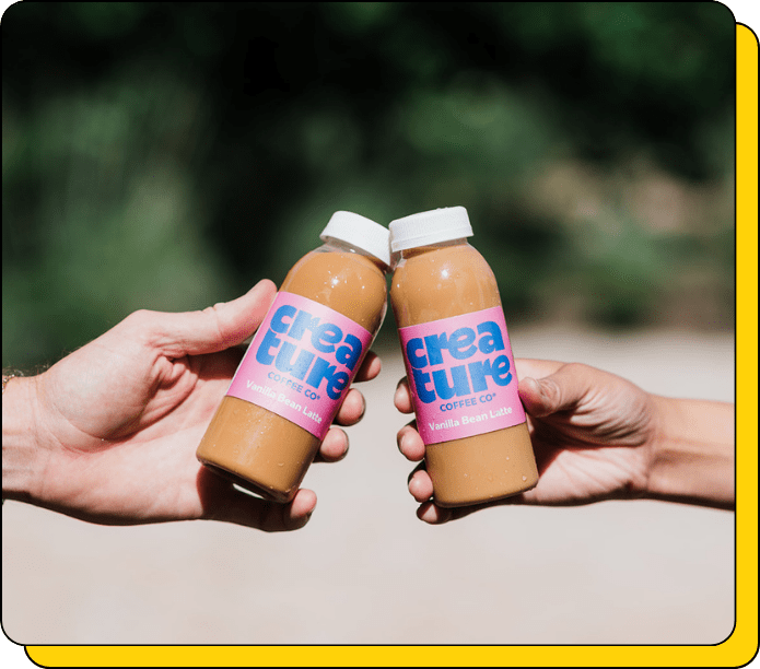 Two hands holding bottles of 'creature coffee co' against a blurred natural background at an event with coffee catering the best Vanilla Bean Lattes
