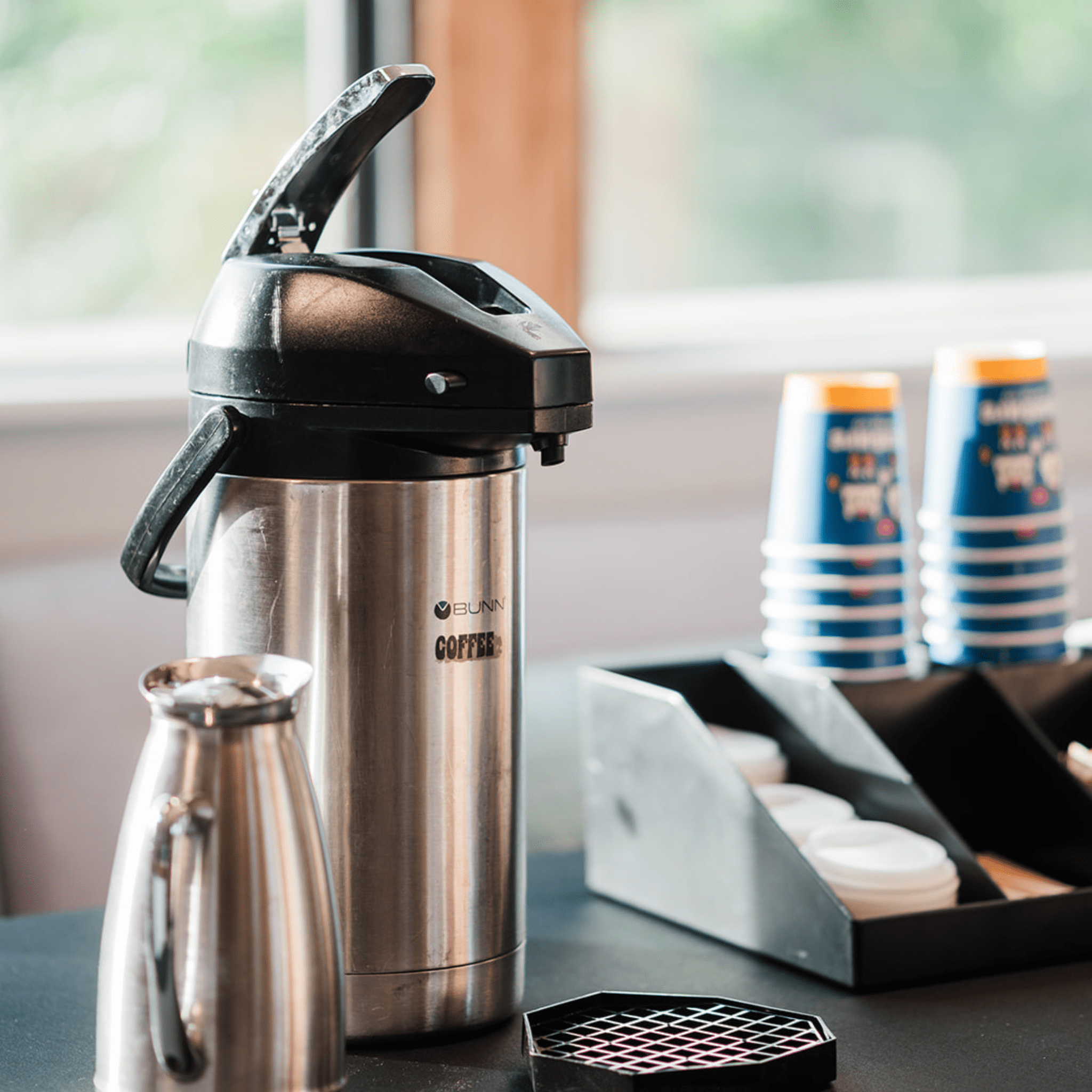 Stainless steel coffee carafe with a black lid on a table with coffee cups and a window in the background with self-service drinks 