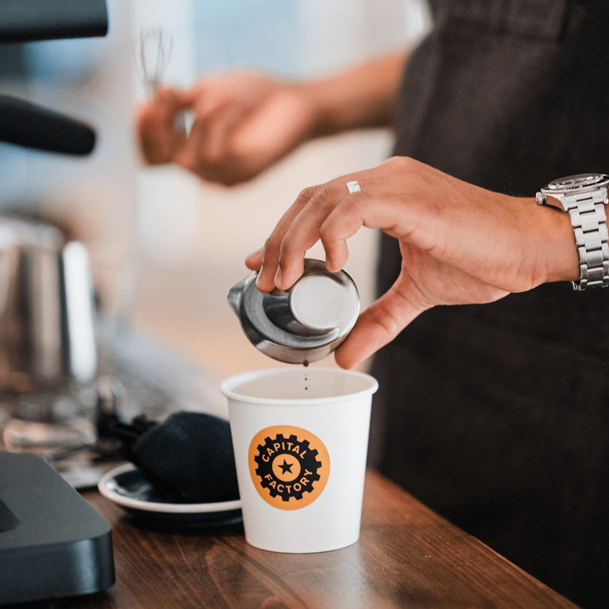 Person pouring coffee into a cup with a visible brand logo on a wooden counter.