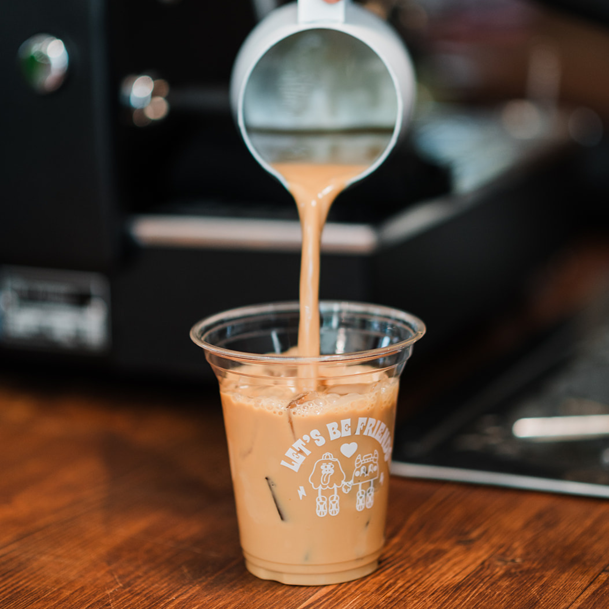 Iced latte being poured into a clear plastic cup with text on a wooden surface.