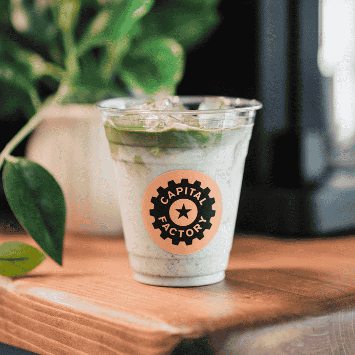 Clear plastic cup with a green drink and 'Capital Factory' logo on a wooden surface with a blurred background