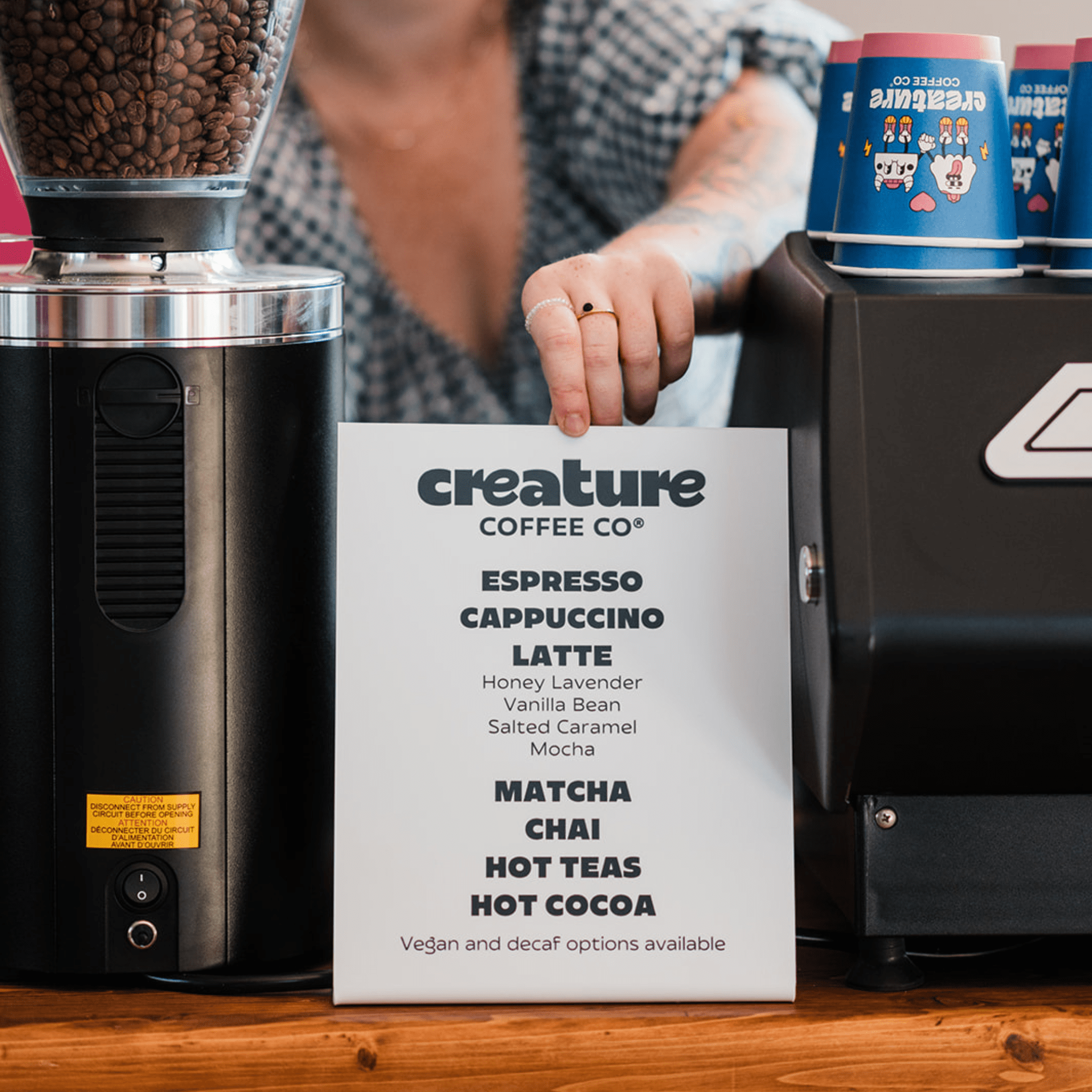 Coffee grinder and coffee machine with a menu for Creature Coffee Co. on a wooden counter.