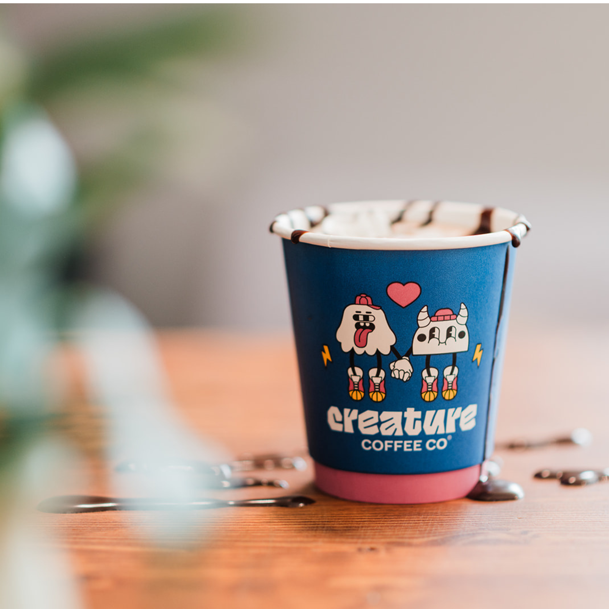 Blue paper cup with hot cocoa and hot chcocolate and 'Creature Coffee Co' branding on a wooden surface.