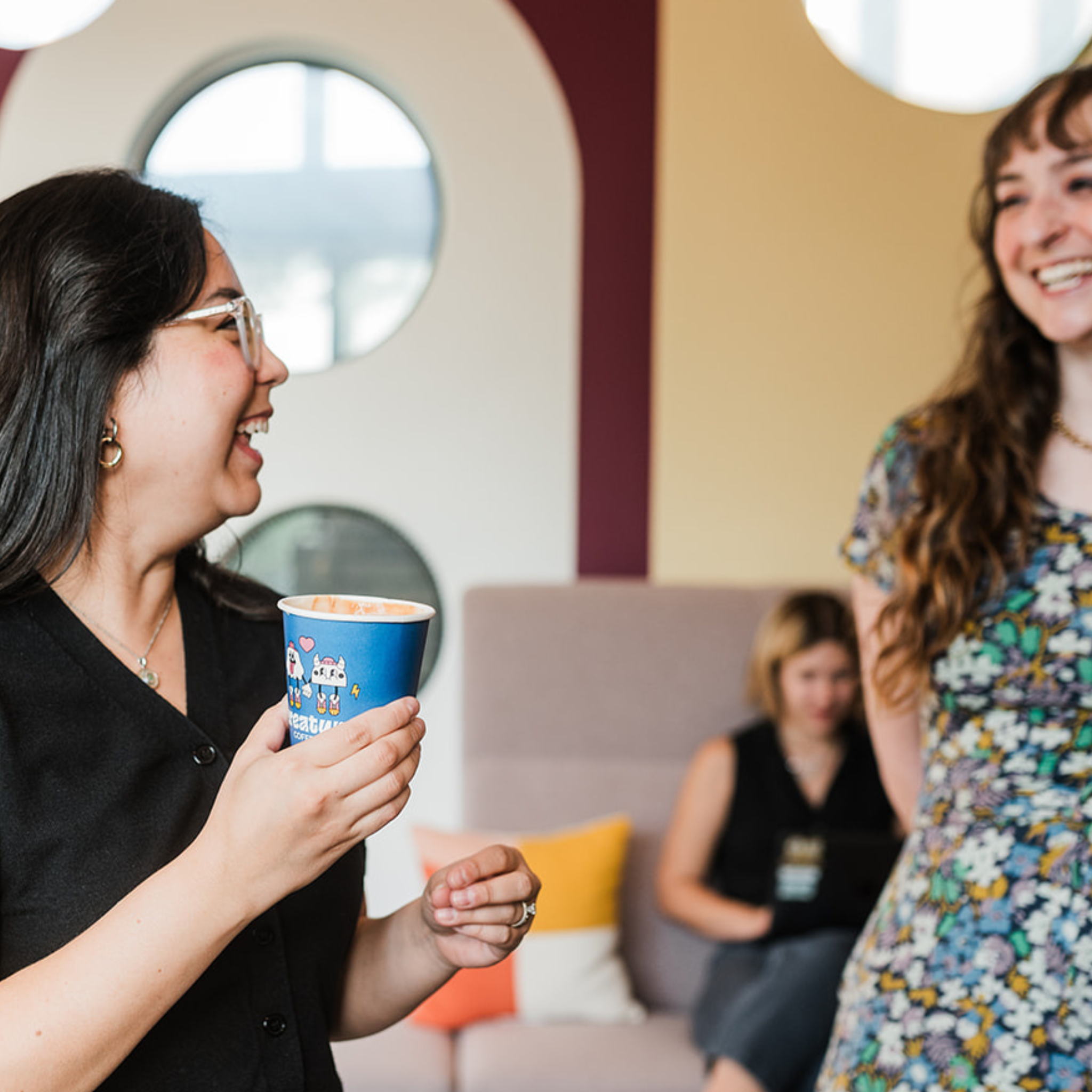 Two women laughing together in a modern indoor setting with a third person in the background.