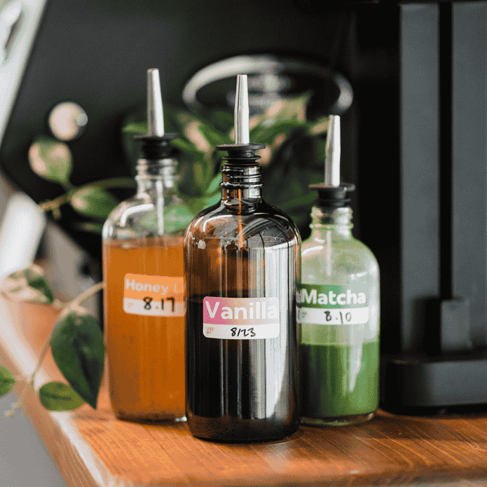 Three bottles labeled 'Honey', 'Vanilla', and 'Matcha' on a wooden surface with a blurred background.
