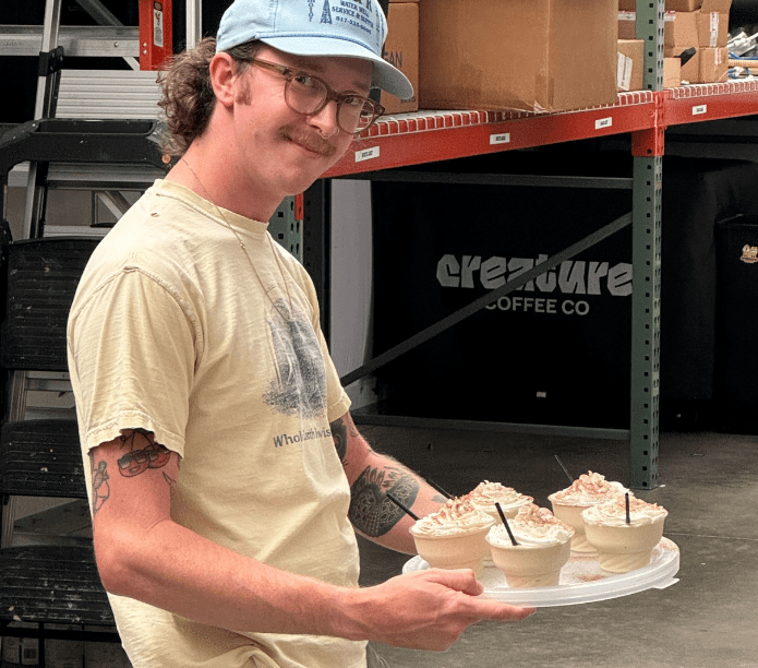 Barista holding a tray of cupcakes in a warehouse setting with 'Creature Coffee Co' branding.