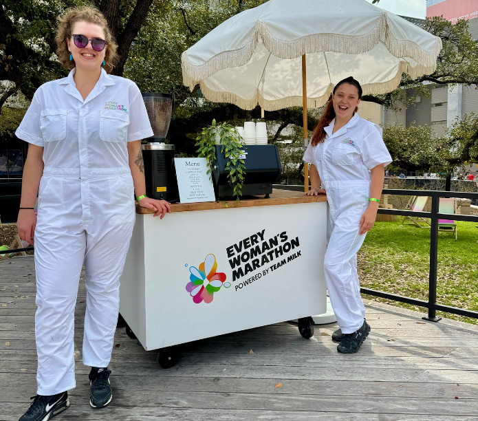 Two women baristas in white custom uniforms standing next to a cart with 'Every Woman's Marathon' branding.
