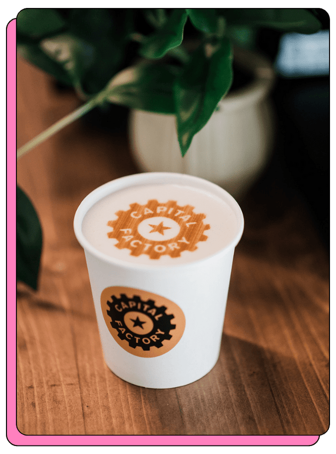 White paper cup with 'Capital Factory' logo printed on top of a latte, on a wooden surface on a mobile coffee cart. Custom-branded coffee cup. 