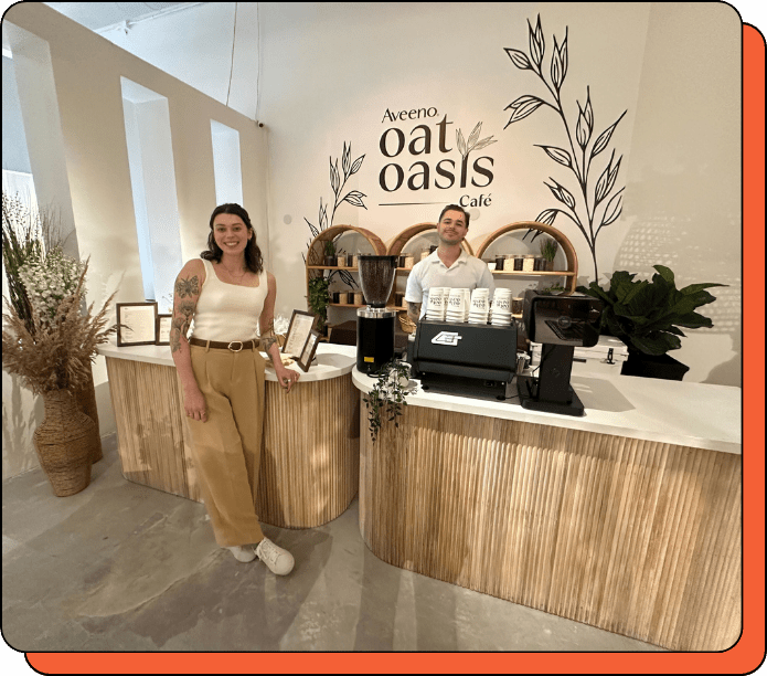 Two people standing in front of a 'Aveeno Oat Oasis Cafe' counter with branding elements. Custom-branded coffee bar from Creature Coffee at an event in SXSW