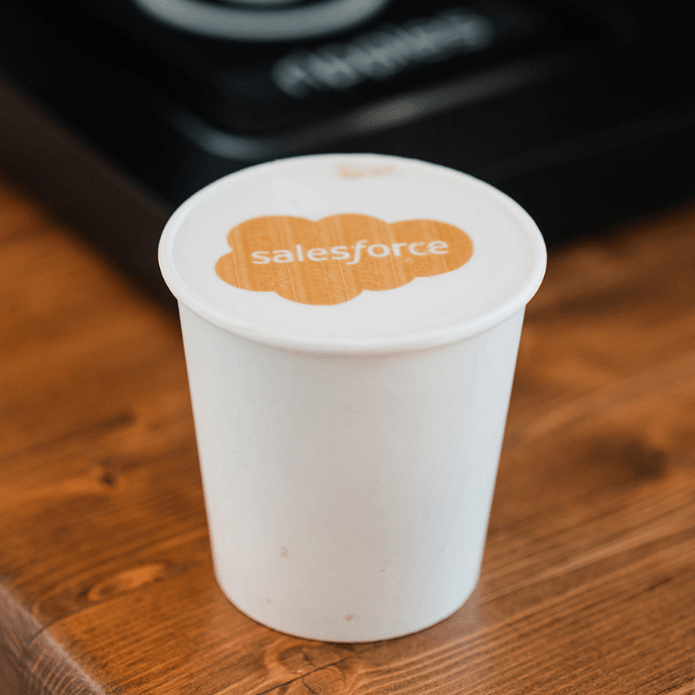White paper cup with Salesforce logo printed on a latte, on mobile coffee cart, on a wooden surface coffee bar top at an event