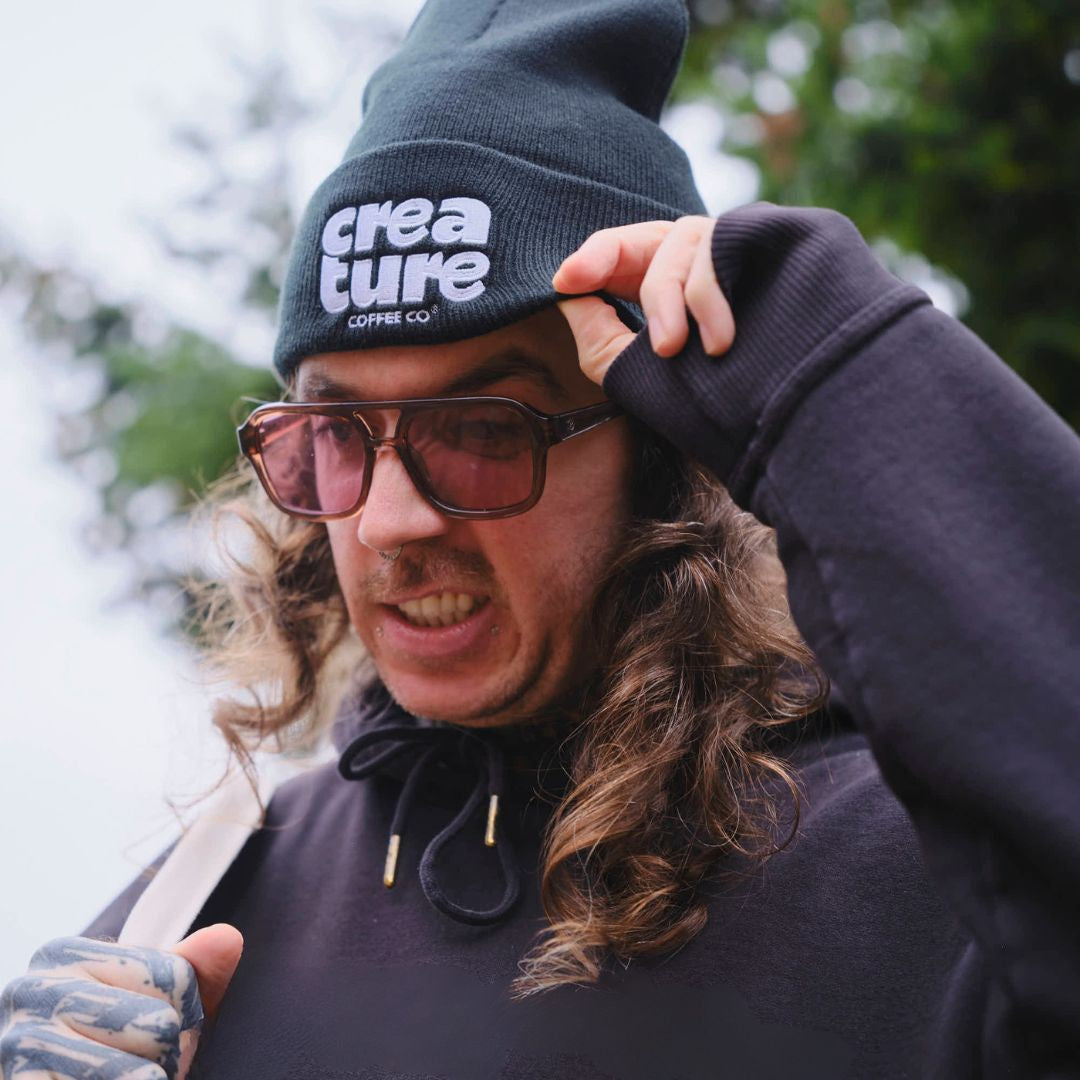 Person wearing a 'creature coffee co' beanie and sunglasses with a blurred natural background. GIft from Specialty Coffee Roaster.