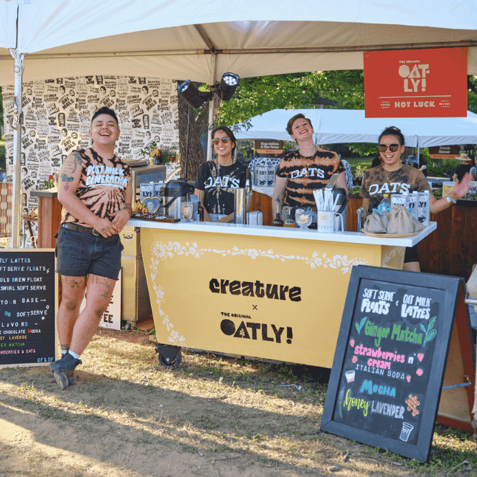 Group of baristaa standing behind a coffee cart stand with Oatly branding at an outdoor event.