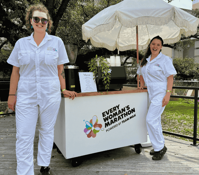 Two women in white uniforms standing next to a coffee cart with 'Every Woman's Marathon' branding outdoors.