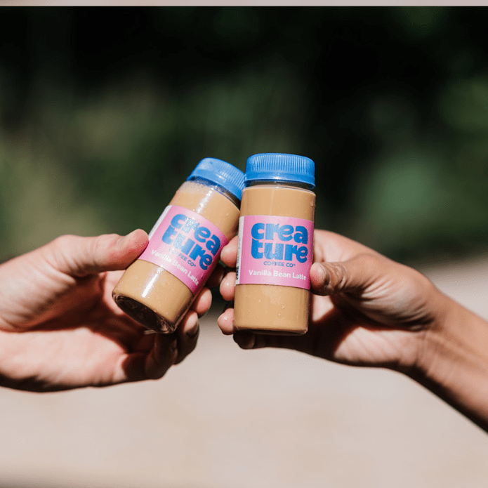 Two hands holding bottled lattes of 'creature' peanut butter against a blurred natural background at a coffee catering event next to a mobile coffee cart. Label reads Vanilla Bean Latte Bottle. 