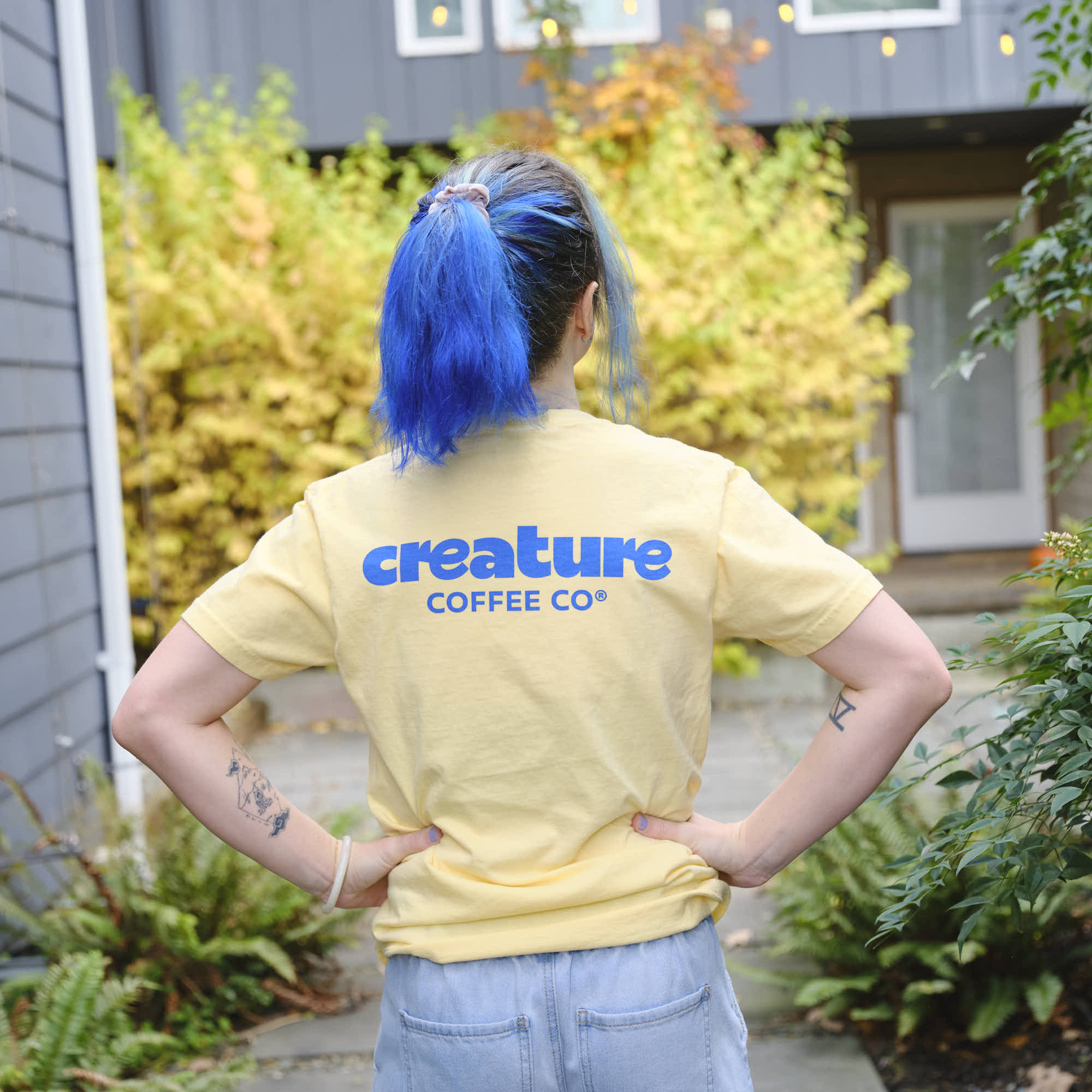 Person wearing a yellow 'creature COFFEE CO' t-shirt outdoors.