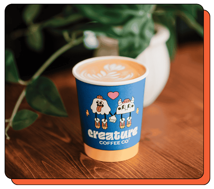 Blue paper compostable creature coffee cup with cartoon characters and 'creature coffee co' branding on a wooden surface. At an event, this cup is served to a client. 