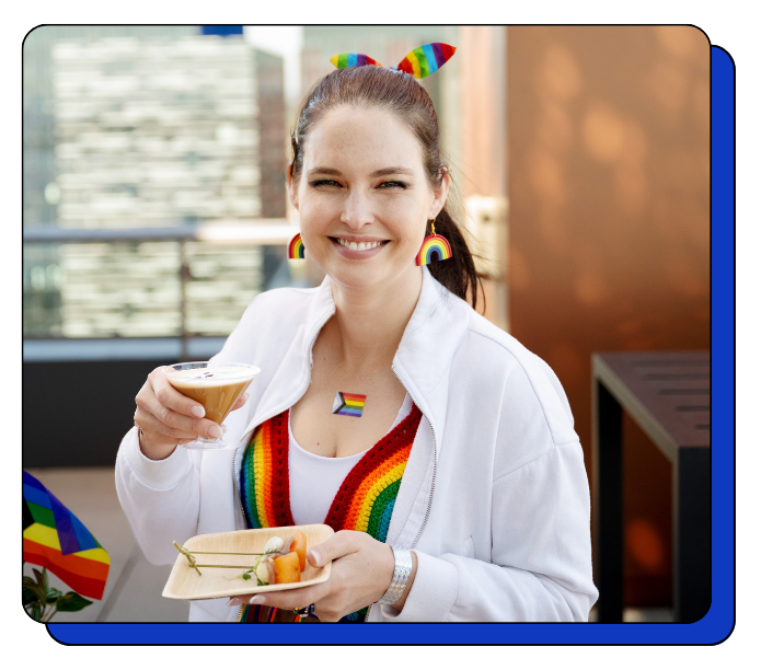 Woman holding a latte drink and snack, at an event with coffee catering, wearing rainbow-themed earrings and a rainbow-colored top, with a cityscape background.